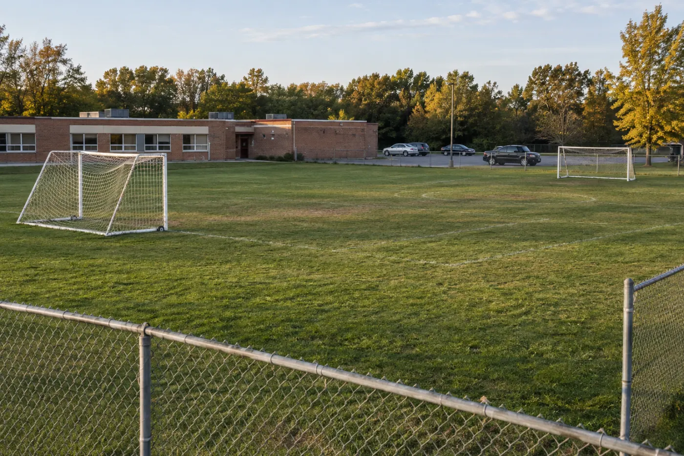 Two small soccer fields on the grass beside a one-storey brick school, a chain-link fence and a parking lot with a few cars