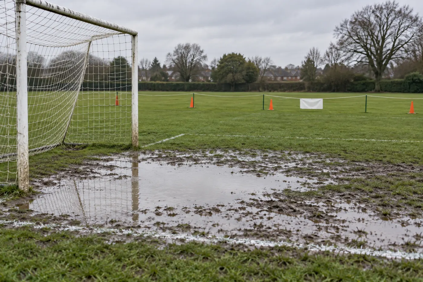 A waterlogged soccer field after overnight rain, standing water in the goal mouth and a rope with orange cones across the touchline