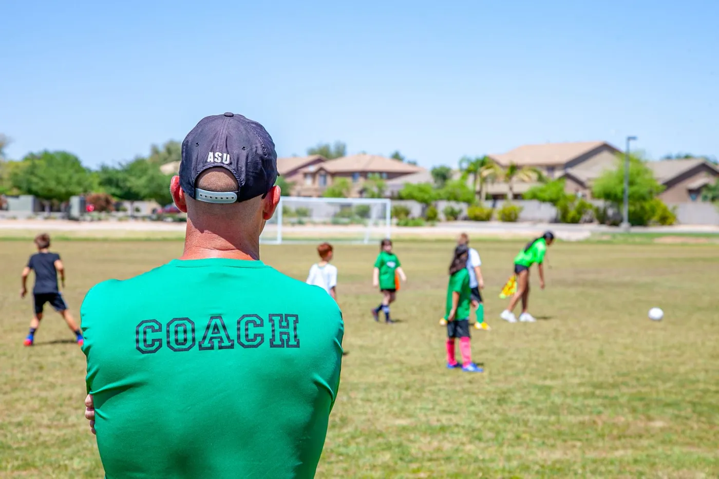 A coach in a cap and a green shirt marked Coach, seen from behind, watching children practise on a sunny field