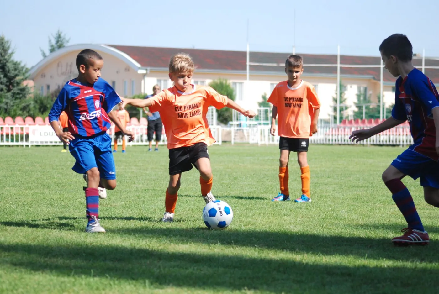 Young players in orange and in blue-and-red kit chasing a ball during a recreational match on a sunny field