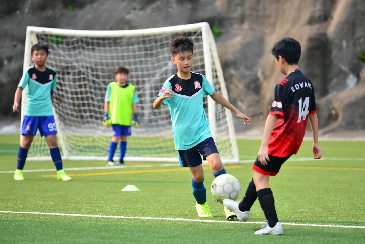 Young players in blue and red kit around a small goal on a sunny field, one about to shoot